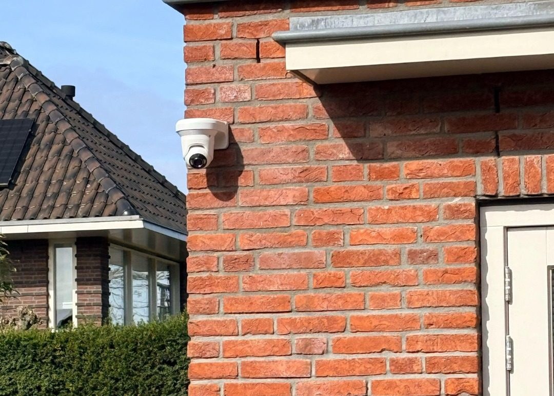 Witte Ajax-camera op de hoek van een bakstenen muur, met een hoge heg erachter