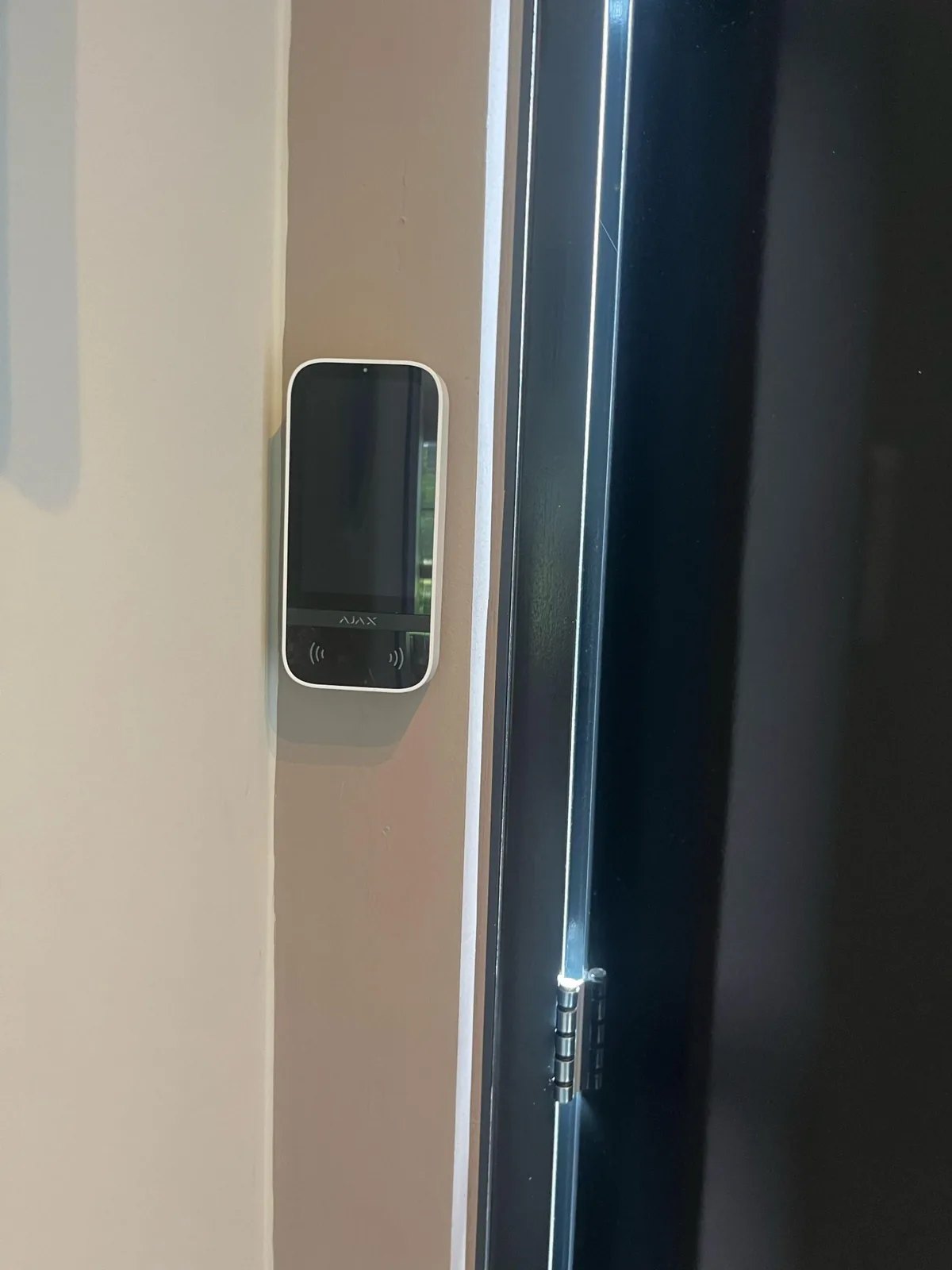 Wit Ajax-bediendeel met touchscreen aan een lichte wand, naast een zwarte deur met scharnieren