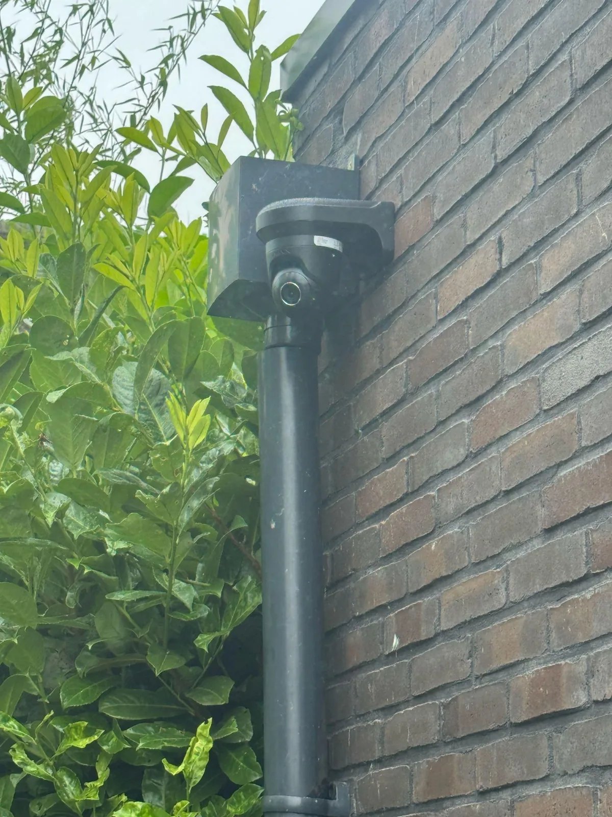 Zwarte camera op de hoek van een bakstenen gevel, met de kabel in een zwarte buis naar beneden langs struiken