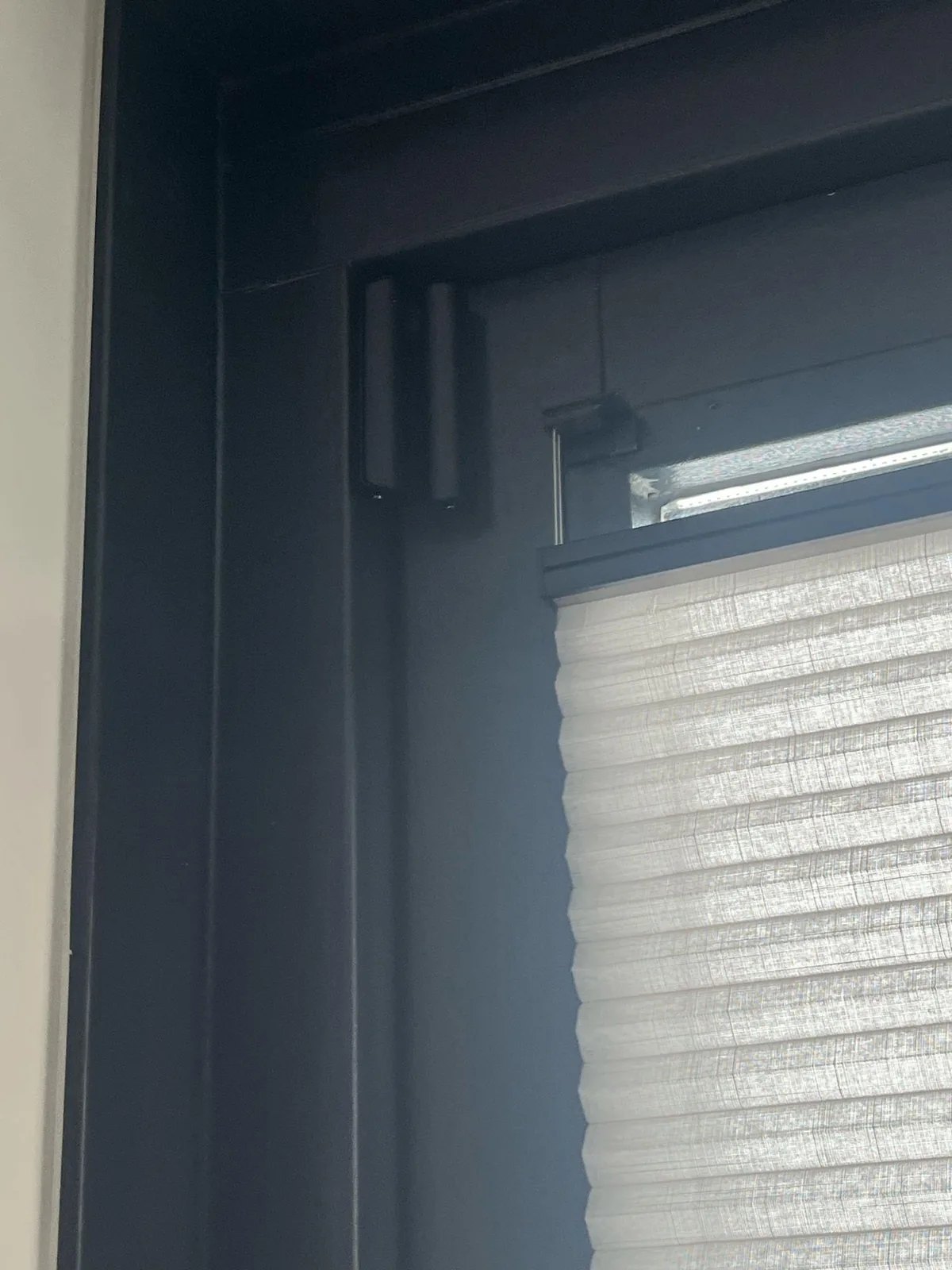Zwart contact in de bovenhoek van een zwart raamkozijn, met een geplooid gordijn eronder
