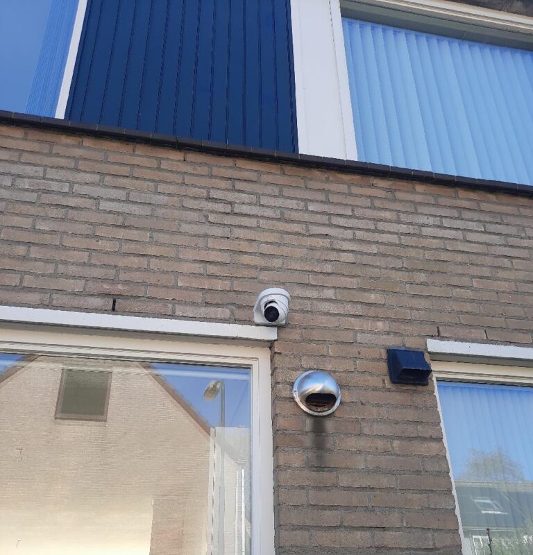 Witte turretcamera op lichtbruin metselwerk, direct boven het kozijn van een glazen pui, naast een rvs-ventilatiekap
