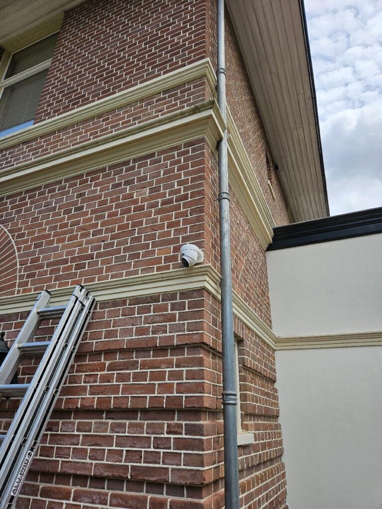 Witte dome-camera op de hoek van een bakstenen gevel, vlak boven een crèmekleurige lijst en naast een zinken regenpijp, met een ladder ertegen