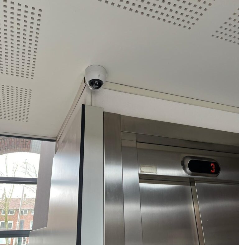 Witte domecamera tegen een geperforeerd plafond, recht boven de roestvrijstalen deuren van een lift met verdiepingsdisplay