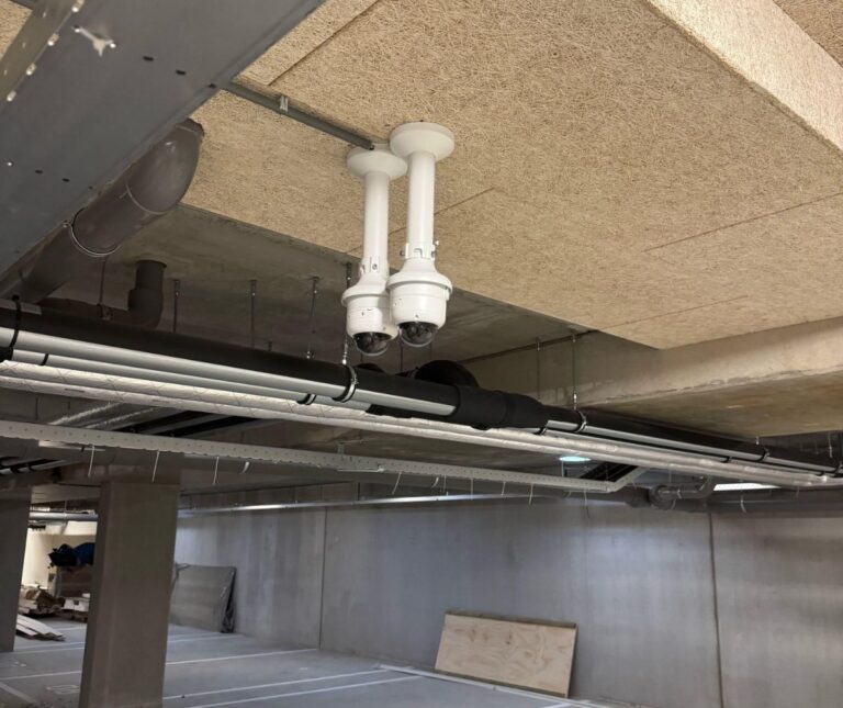 Twee witte domecamera's op verlengbuizen aan het plafond van een parkeergarage, tussen leidingen en betonnen kolommen