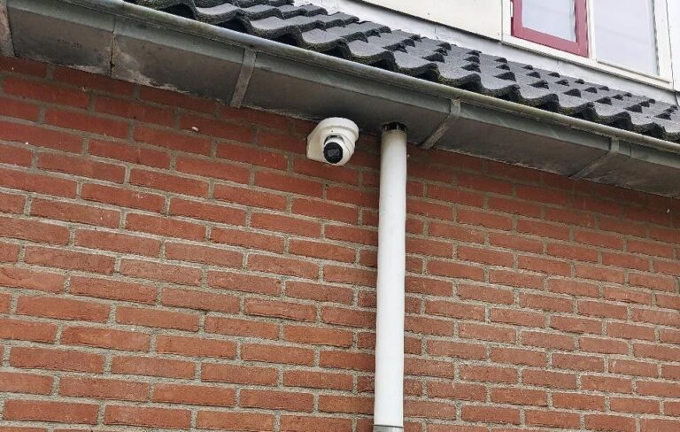 Witte turretcamera onder een grijze dakgoot op rode baksteen, direct naast een witte regenpijp