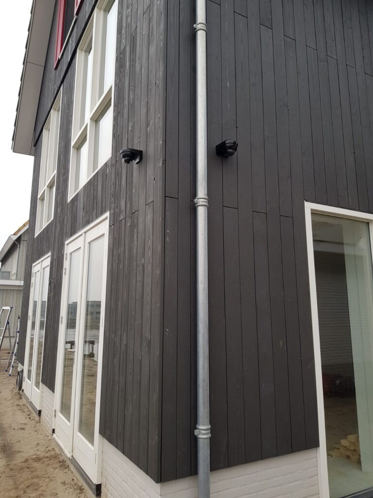 Twee zwarte camera's op de hoek van een woning met zwarte verticale houten gevel, naast een zinken regenpijp en witte kozijnen