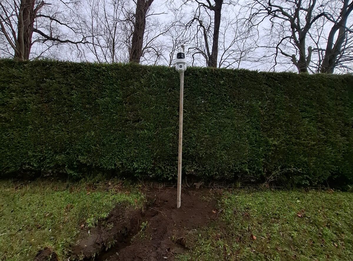Witte camera op een paal voor een hoge groene haag, met een verse sleuf in het gras ernaartoe