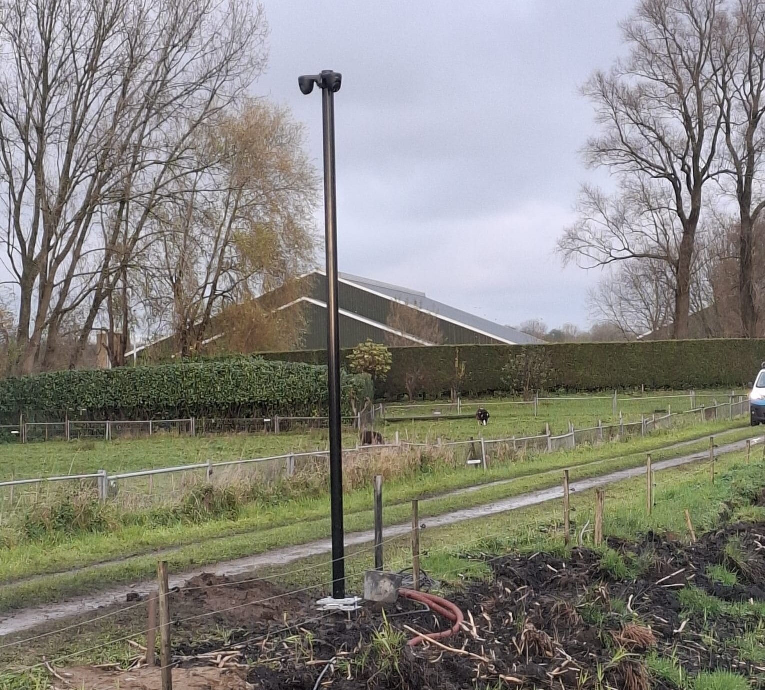 Zwarte mast met camera op een voetplaat aan de rand van een weiland, met een rode kabelbuis bij de voet