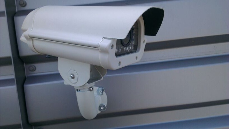 Witte camera in een behuizing met zonnekap op een verstelbare beugel tegen een grijze geprofileerde wand