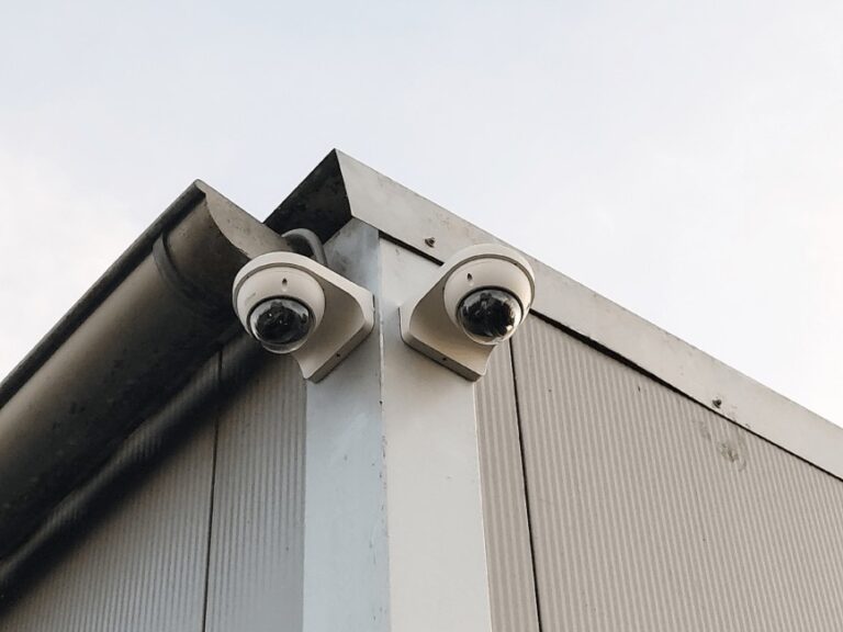 Twee witte domecamera's op hoekbeugels bovenaan de hoek van een gebouw met grijze plaatbekleding