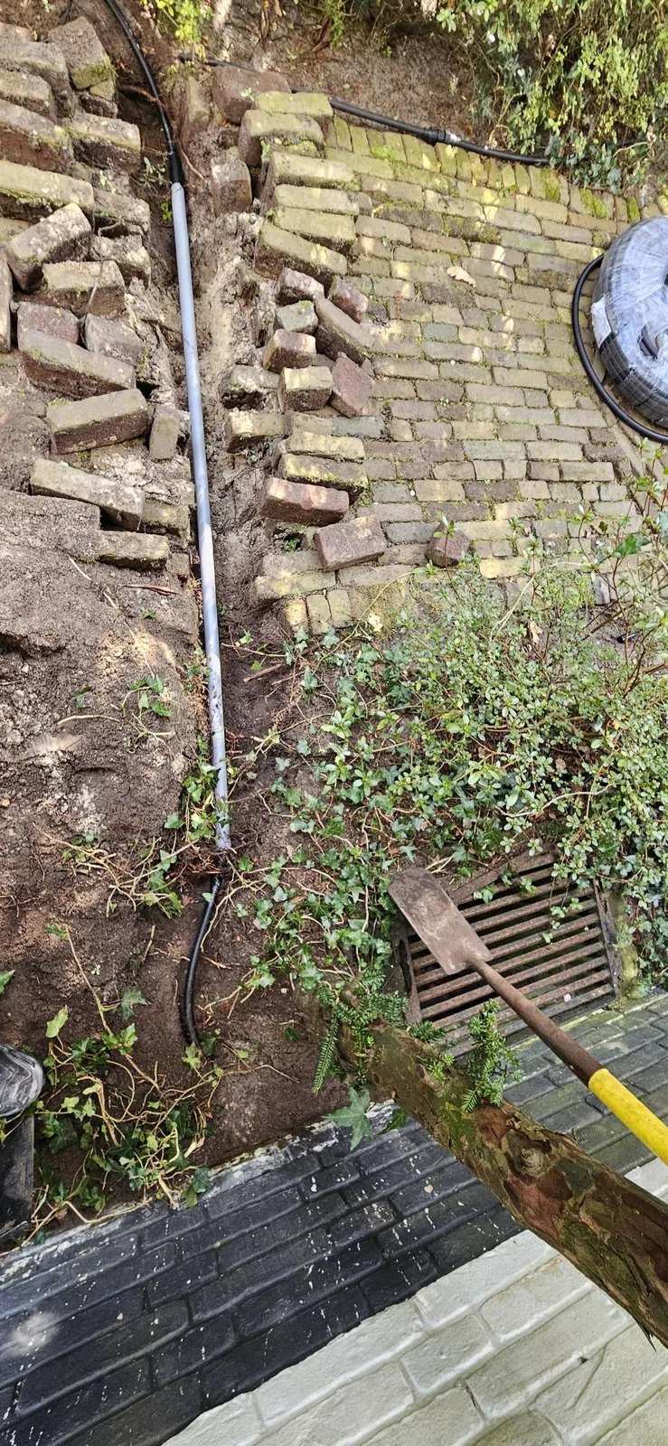Opengegraven sleuf in de tuin met een grijze buis die onder een bakstenen looppad door loopt, losse klinkers ernaast