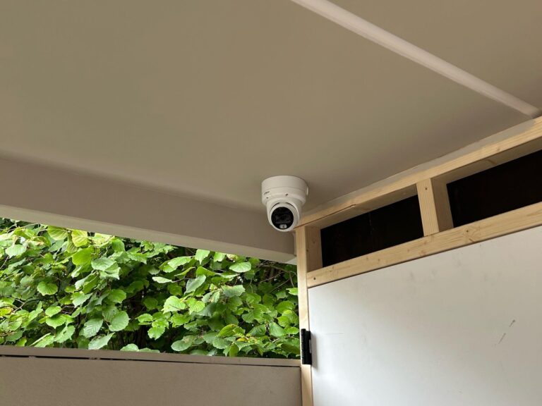 Witte camera aan het plafond van een overkapping, met groene haag op de achtergrond