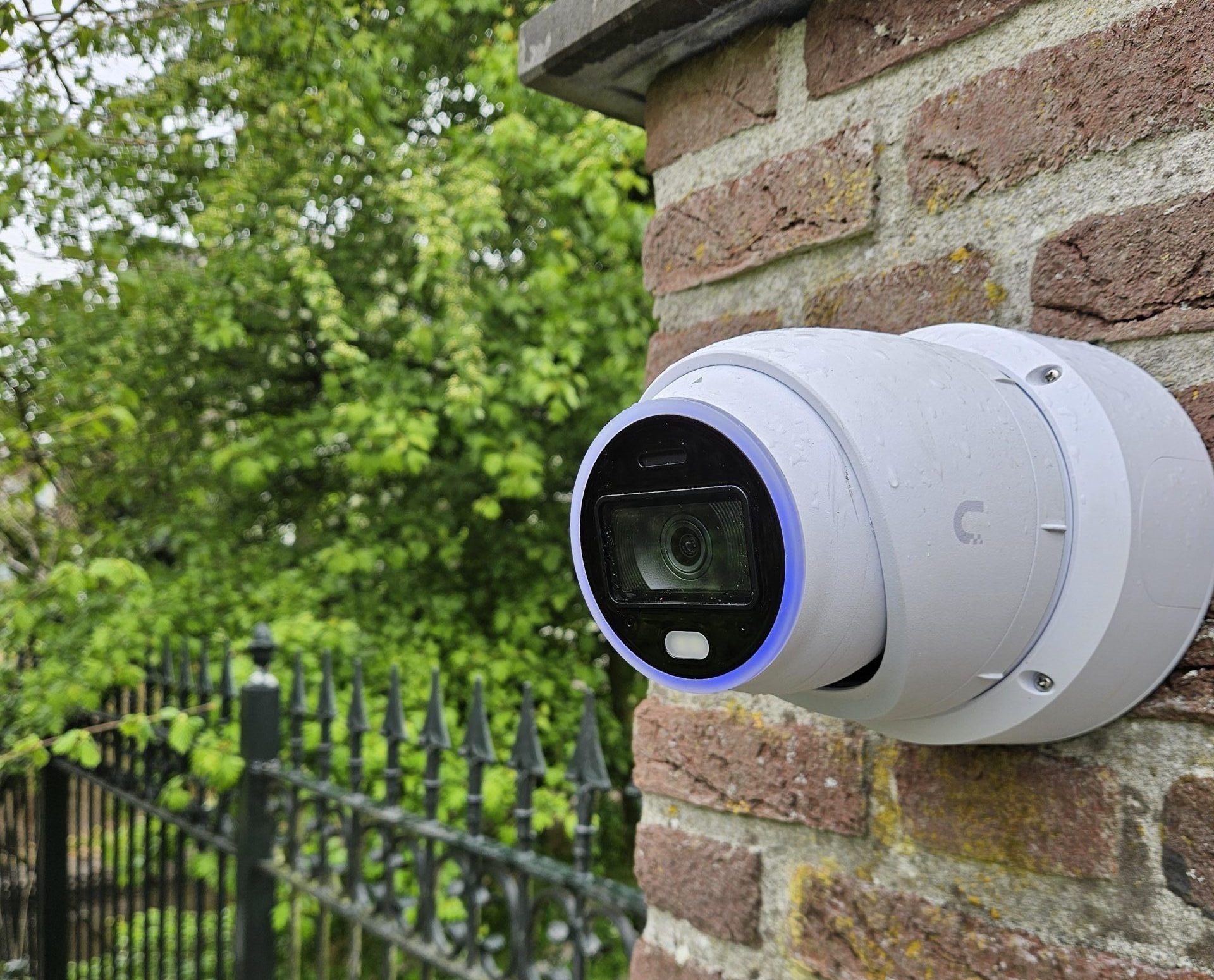 Witte camera op een bakstenen pilaar naast een zwart smeedijzeren hek met groen erachter