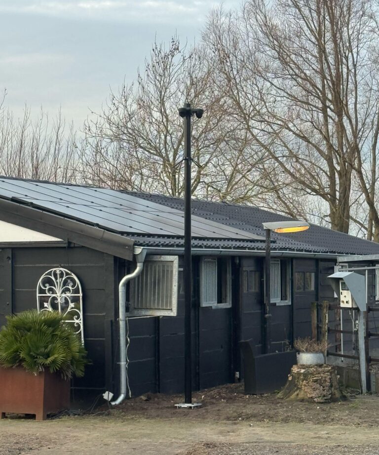 Zwarte mast naast een zwarte paardenstal met zonnepanelen op het dak en kale bomen erachter