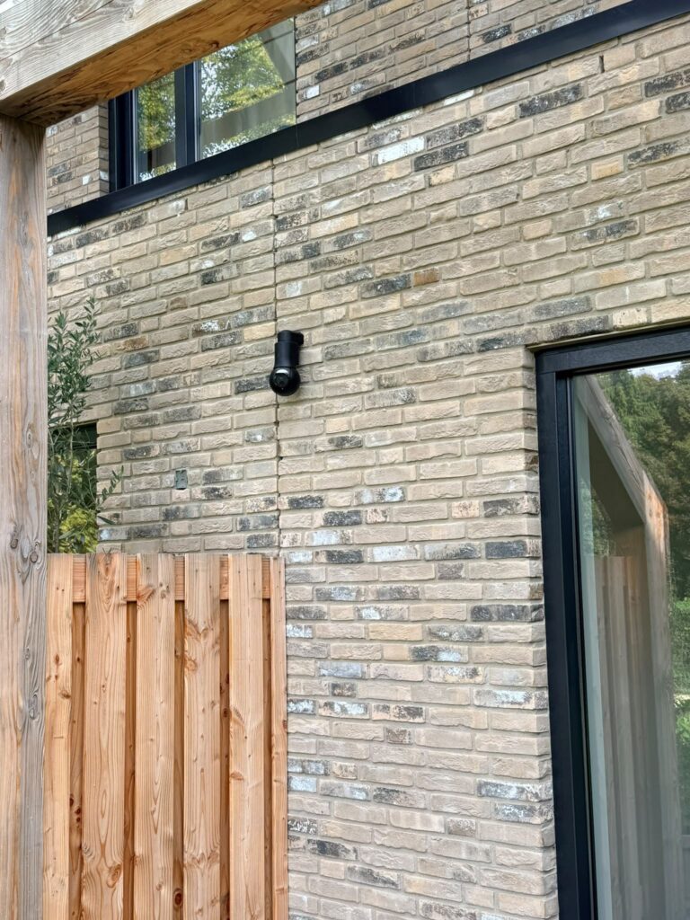 Zwarte draaibare camera op een lichte bakstenen gevel boven een houten schutting, naast een zwart kozijn