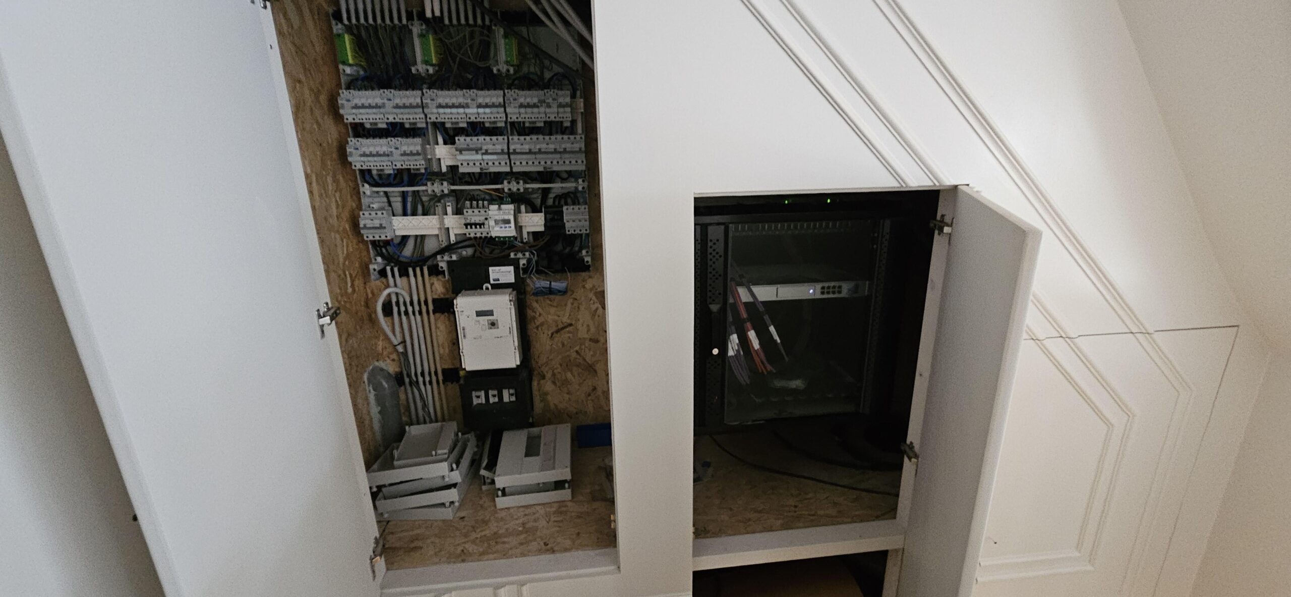 Witte inbouwkast onder een schuine wand, links de open meterkast, rechts een zwart netwerkrack