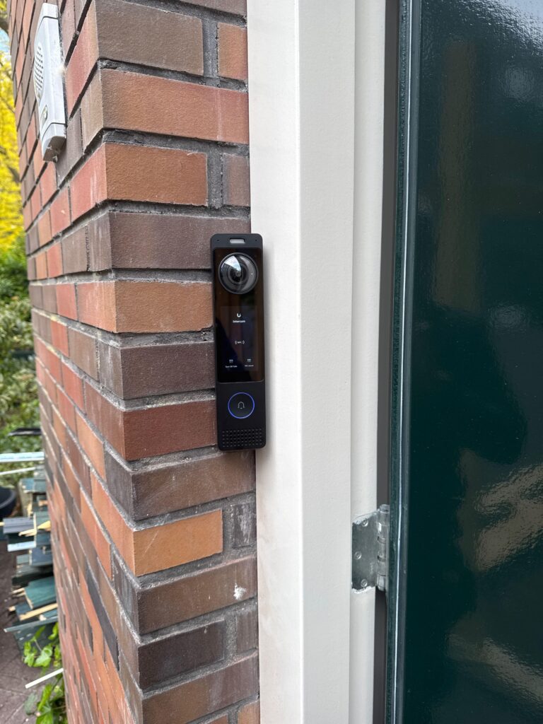 Smal zwart toegangspunt met lens en knoppen op het metselwerk naast het kozijn van een donkergroene voordeur