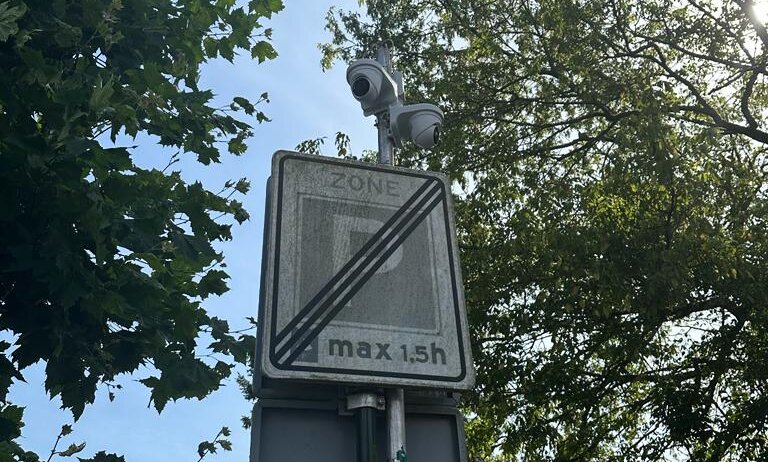 Twee witte camera's bovenop een paal met een bord einde parkeerzone, tegen bomen en een blauwe lucht