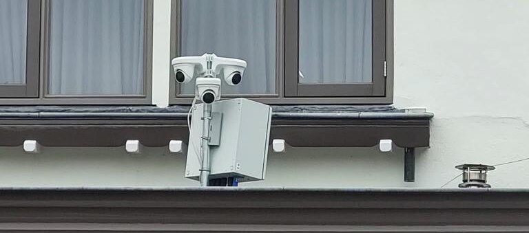Drie witte camera's op een korte buis met een grijze systeemkast eronder, gemonteerd op een gevelrand onder twee ramen