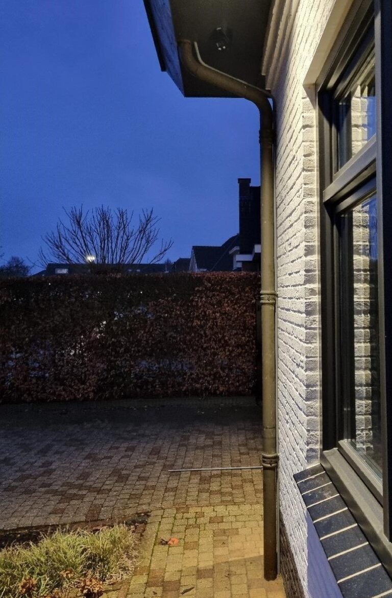 Witte gevel bij schemer met een donkere regenpijp; bovenin zit een zwarte camera in het donkere boeideel, met een oprit en een beukenhaag erachter