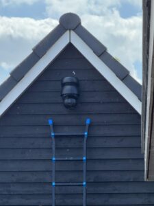 Zwarte draai- en zoomcamera hoog in de punt van een donkere houten schuurgevel, met een ladder eronder