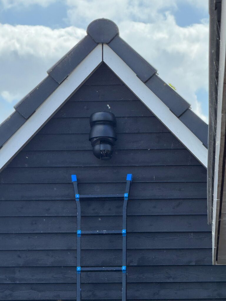 Zwarte draai- en zoomcamera hoog in de punt van een donkere houten schuurgevel, met een ladder eronder