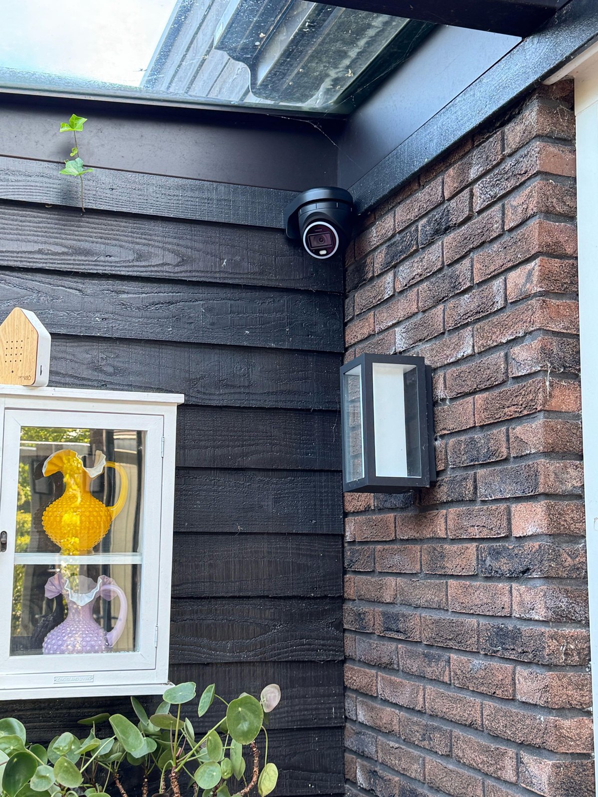 Zwarte turretcamera in de hoek van een overkapping, tussen donker hout en baksteen, boven een buitenlamp