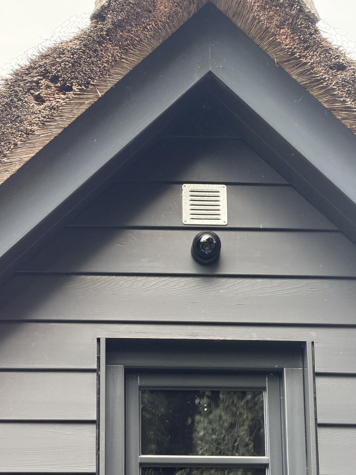 Kleine zwarte camera op een donkergrijze houten gevel onder een rieten kap, vlak onder een ventilatierooster