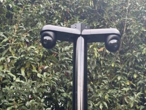 Zwarte tuinmast met bovenop twee camera's die elk een andere kant op kijken, voor een groene haag