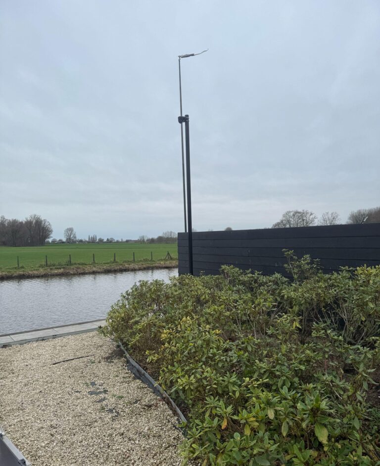 Zwarte cameramast achter in een tuin met heesters, naast een sloot met weiland erachter