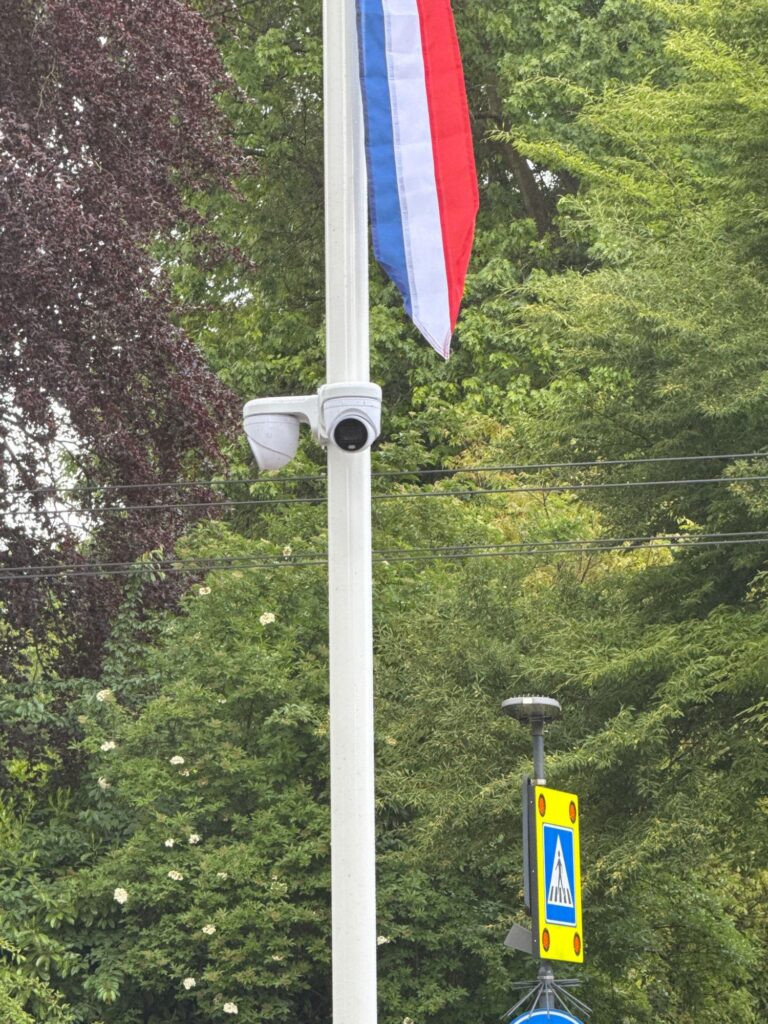 Twee witte camera's op een beugel aan een witte vlaggenmast, met de Nederlandse vlag erboven en bomen erachter