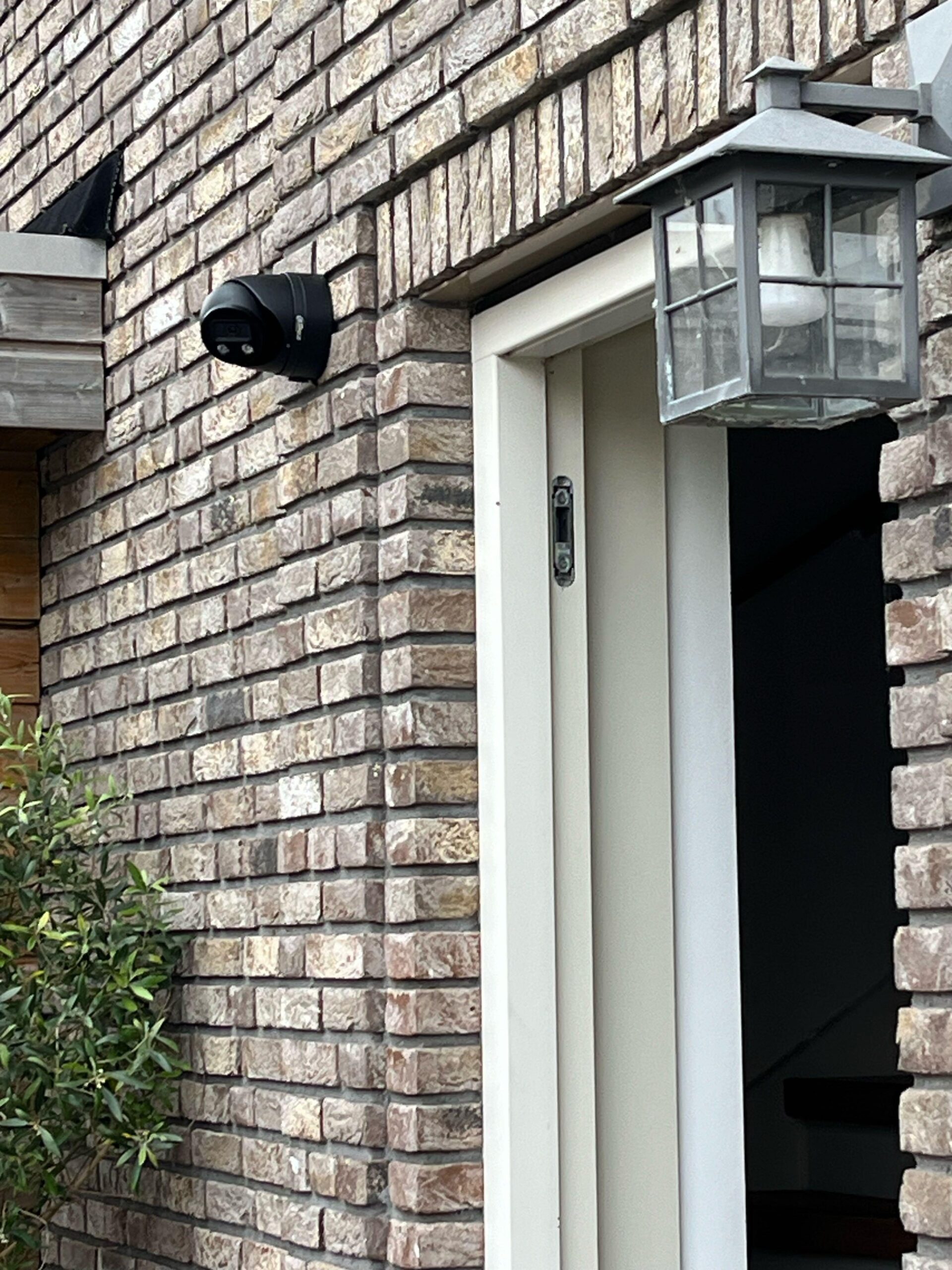 Zwarte camera op het metselwerk naast een voordeur, met een buitenlamp aan de andere kant van de deur