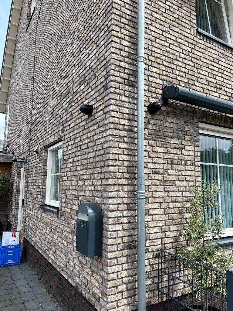 Twee zwarte camera's hoog op de hoek van een bakstenen gevel, naast een regenpijp en een antraciet brievenbus