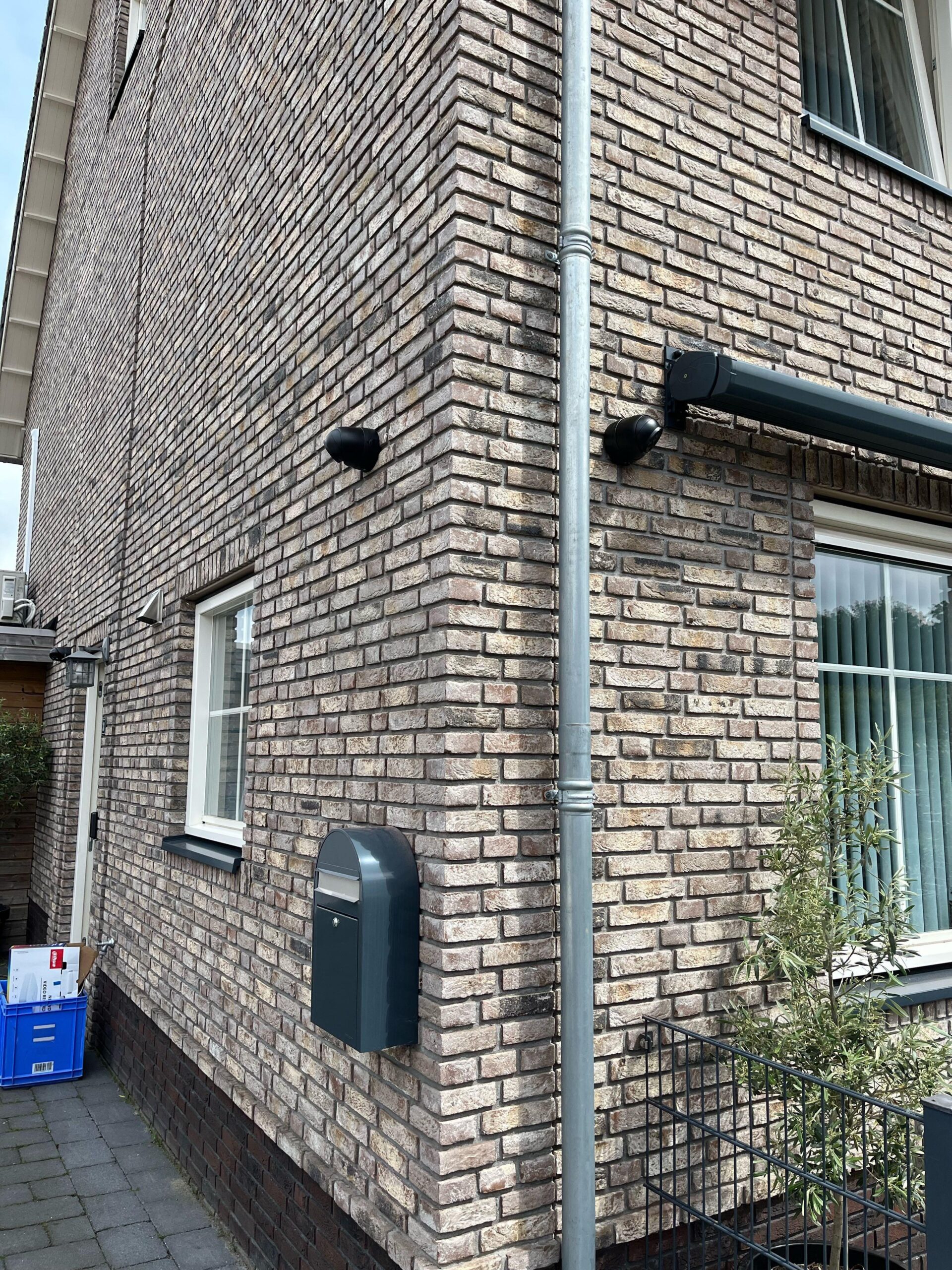 Twee zwarte camera's hoog op de hoek van een bakstenen gevel, naast een regenpijp en een antraciet brievenbus