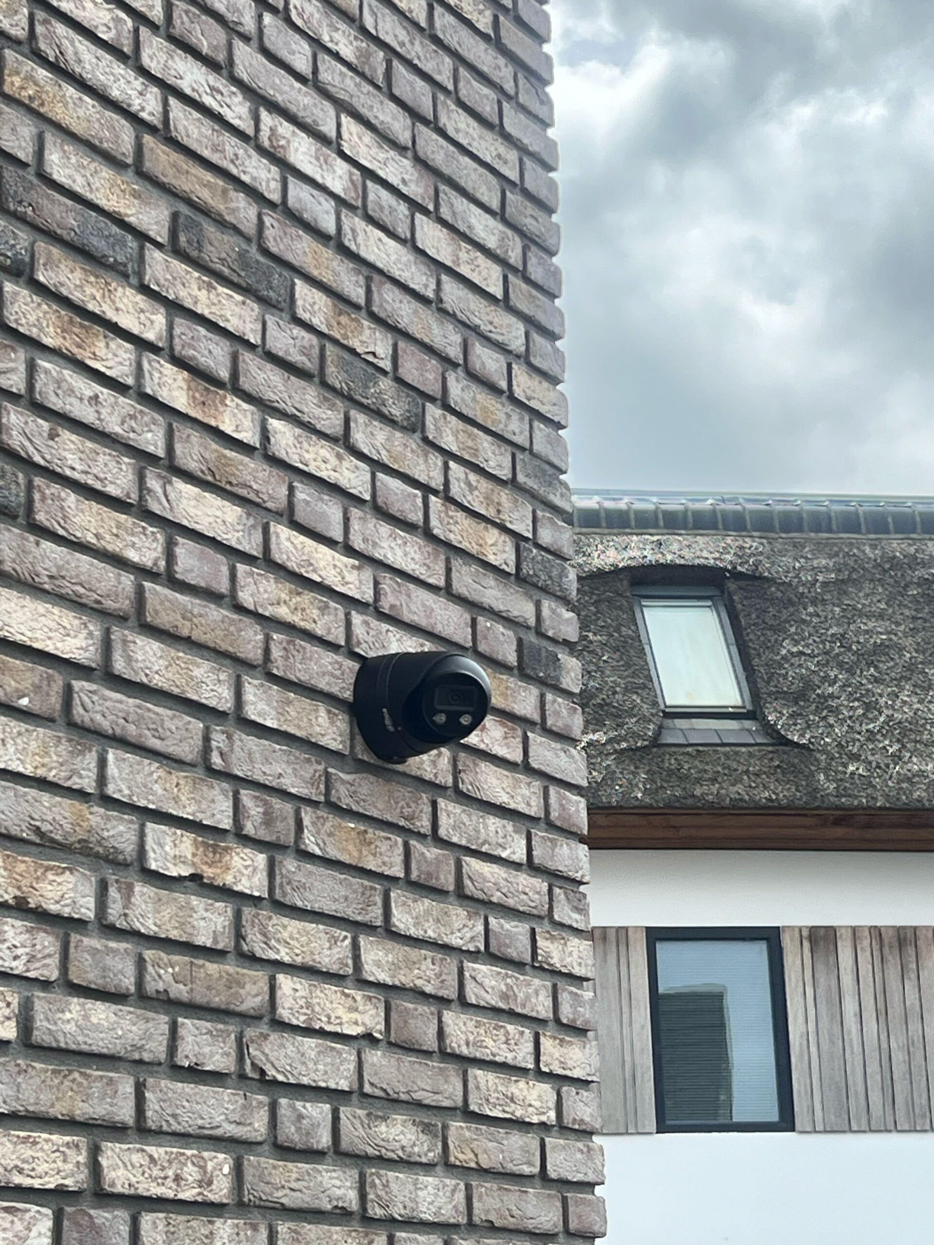 Zwarte camera op een bakstenen muur, met daarachter een buurpand met rieten kap